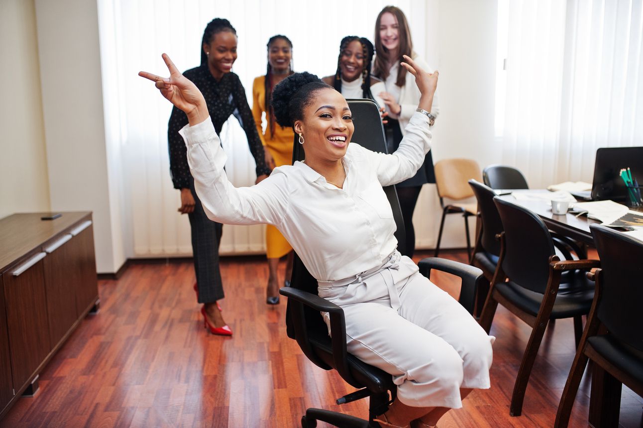Five multiracial business womans standing office roll woman chair diverse group female employees formal wear having fun